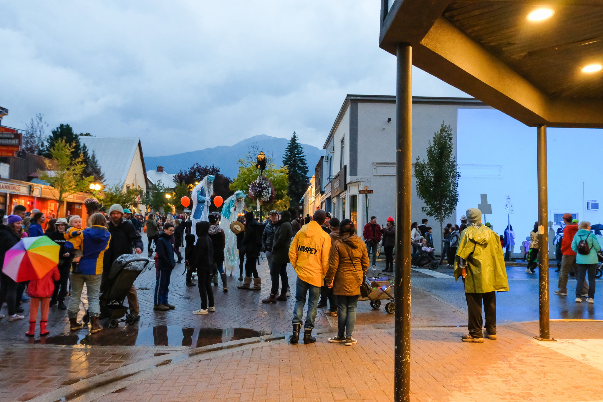 A crowd of people at sunset gathered in a downtown square interacting with each other and theatrical performers. 