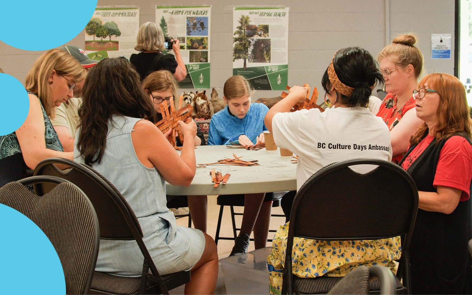 A group of individuals seated at a table working on cedar woven hearts. An Indigenous woman facilitator leads the group as they create their artworks.
