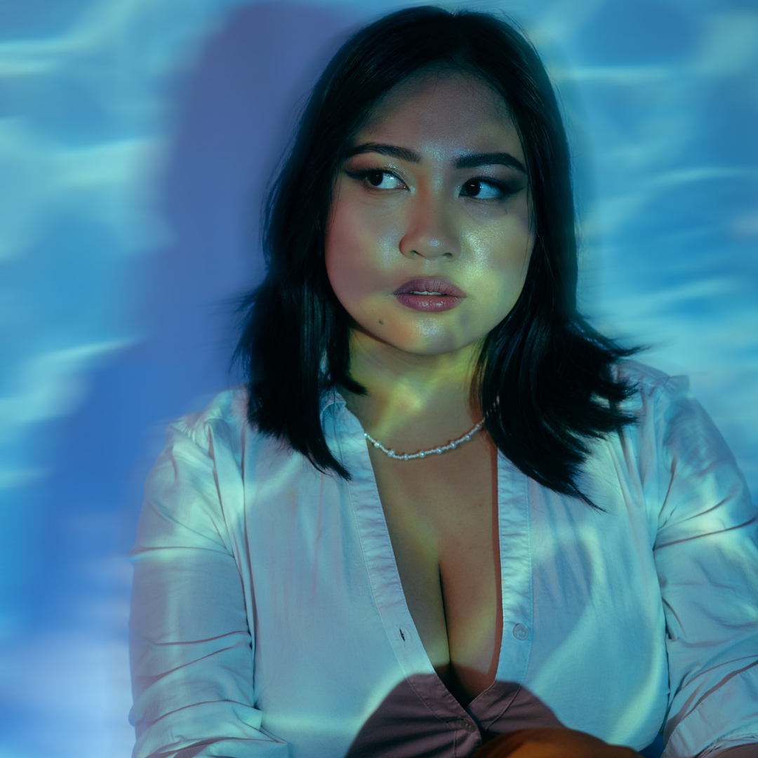 A person with black shoulder length hair posing for the camera while looking off in the distance. They are wearing a long white blouse with a deep v-beck and a pearly necklace.