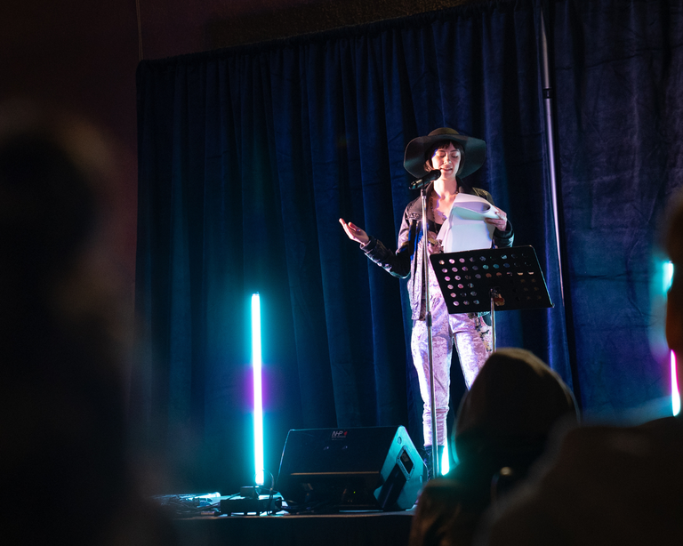 A performer stands with a microphone on a dark stage that is illuminated with colourful lights.
