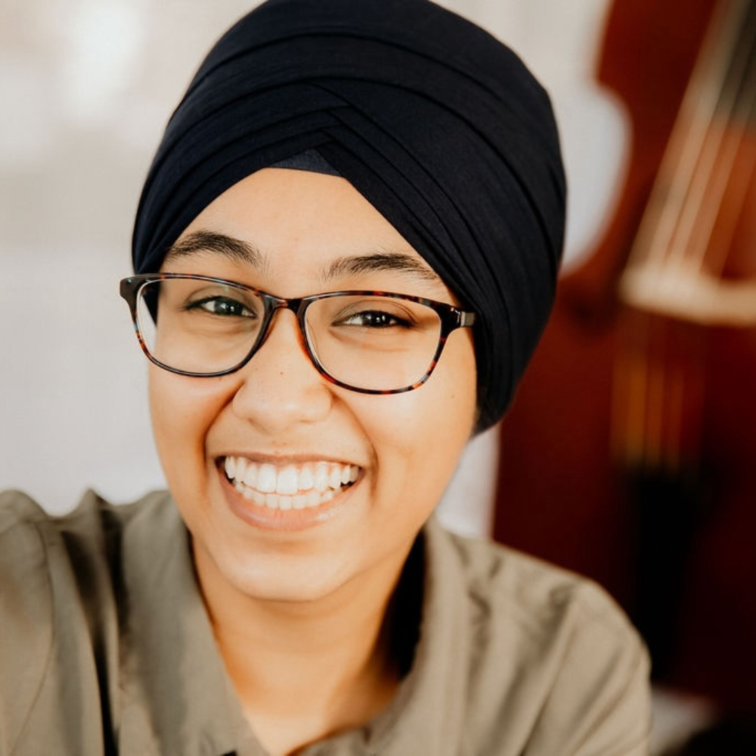 Woman smiling brightly to the camera. She is wearing a black turban, brown glasses, and a khaki shirt.