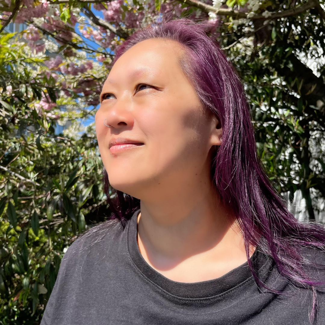 A person posing for a portrait looking off into the distance. Their hair is long and purple and they are smiling softly with a profile view. 