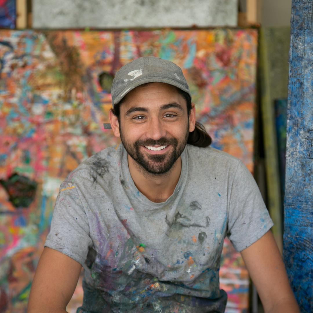 Man smiling at the camera in front of a bright, colourful abstract painting. He has a full beard and long dark hair pinned back in a ponytail. Wearing a grey t-shirt covered in paint.