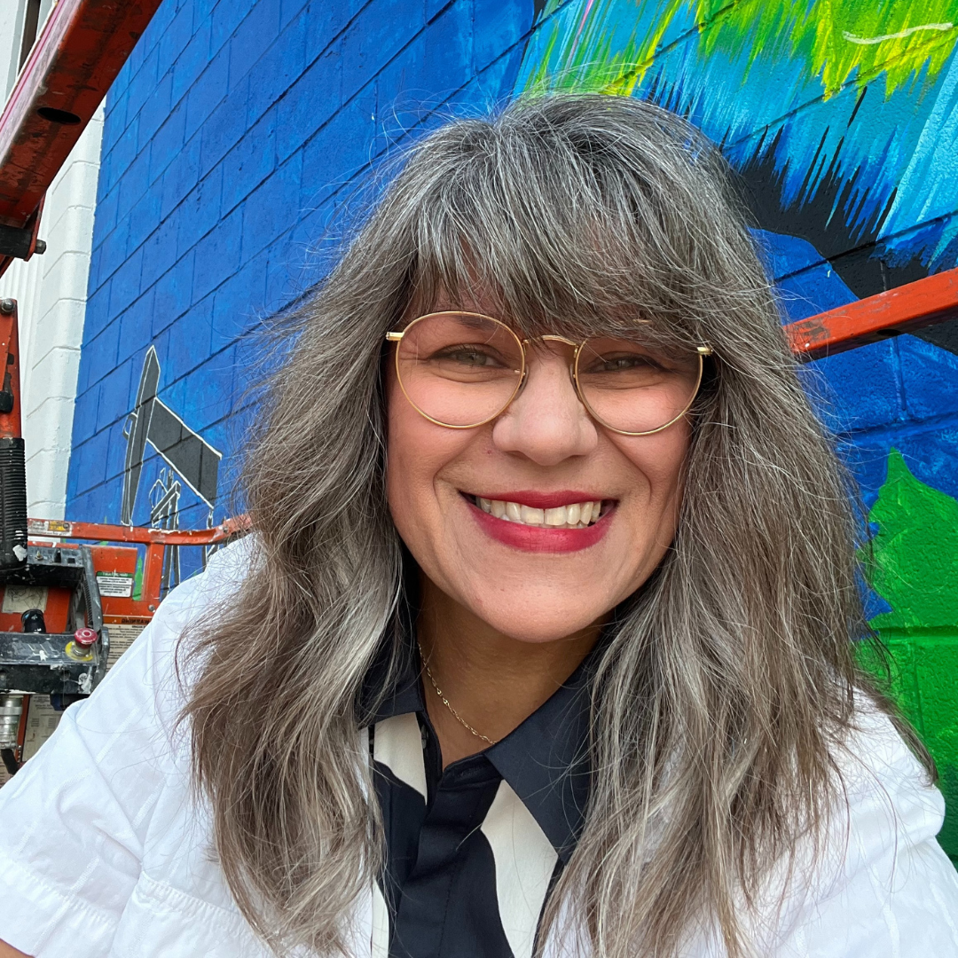 A woman smiling for a photo in front of a colourful outdoor mural. She has large round wire glasses and mid-length grey hair with full bangs. 