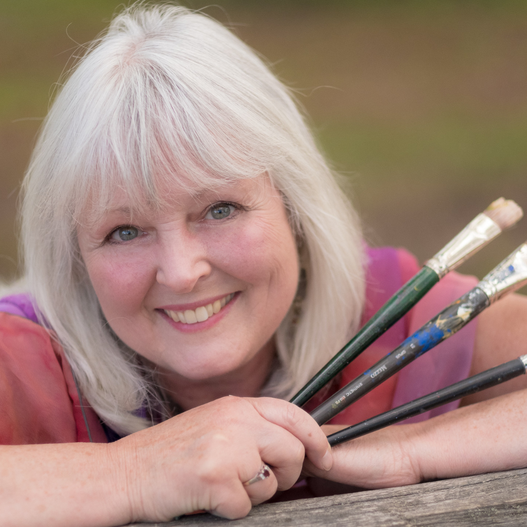 A woman with short grey hair smiling while posing with three paintbrushes in her hand. 