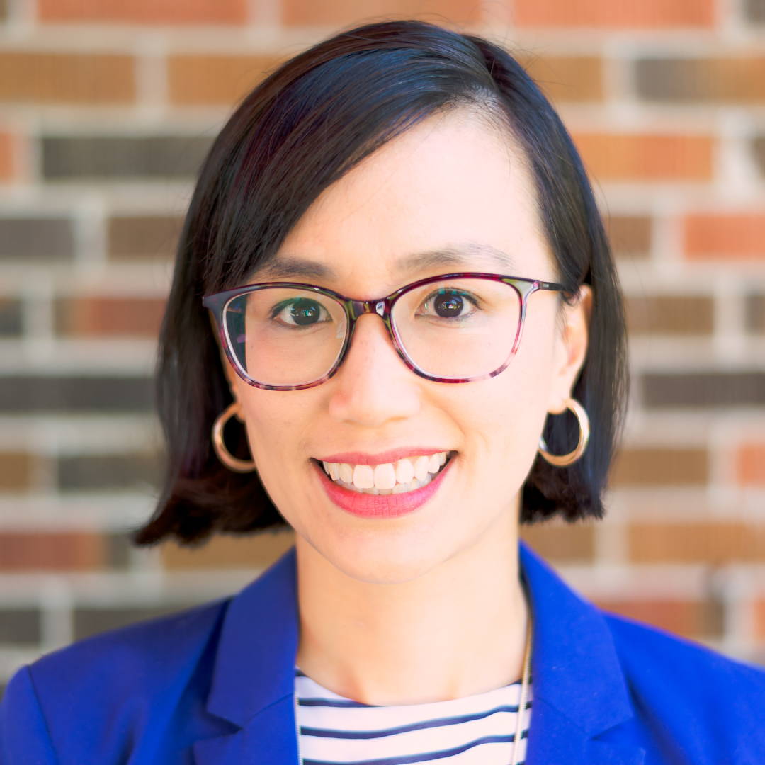 A straight on headshot of a woman with short black hair. She's wearing a bright blue blazer, gold hoop earrings, and brown acetate glasses.