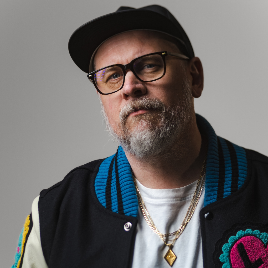 A headshot of a man wearing a vintage-style jacket with colourful patches, a baseball cap, and gold jewelry. He has a full grey beard and black square glasses.