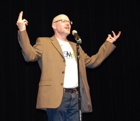 A stand-up comic shown on a stage with his hands in the air. He's bald and is wearing classes, a brown blazer, and a t-shirt that says SMH.