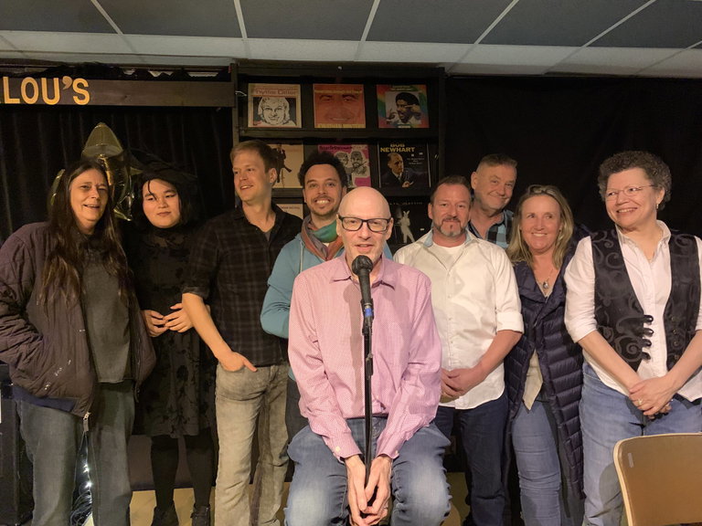 A group of men and women smiling for a group photo. They are in a comedy club and the man at the centre of the image is sitting in front of a microphone.