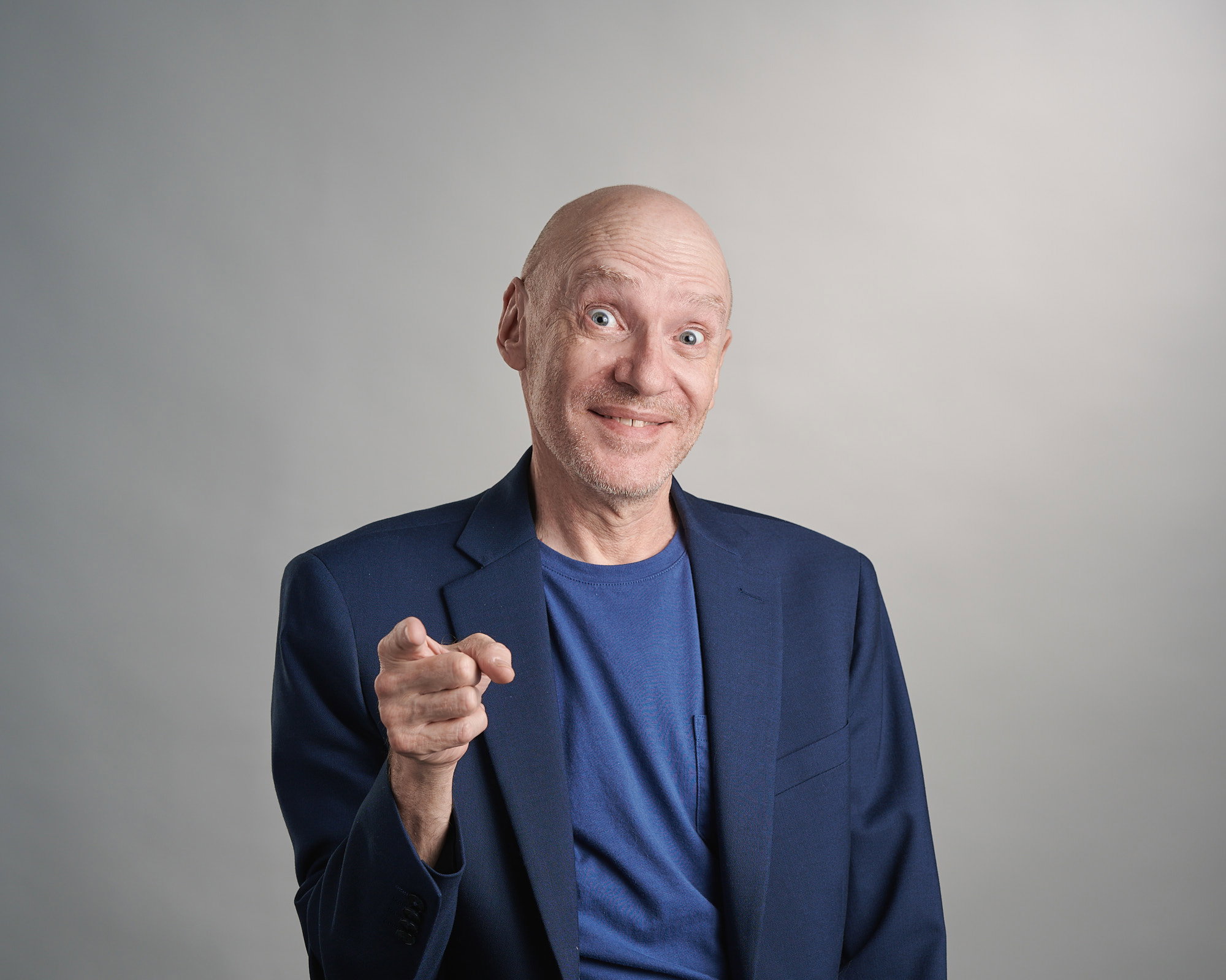 A bald man pointing at the camera while posed in front of a grey backdrop. He's smiling with his brow and eyes wide open.