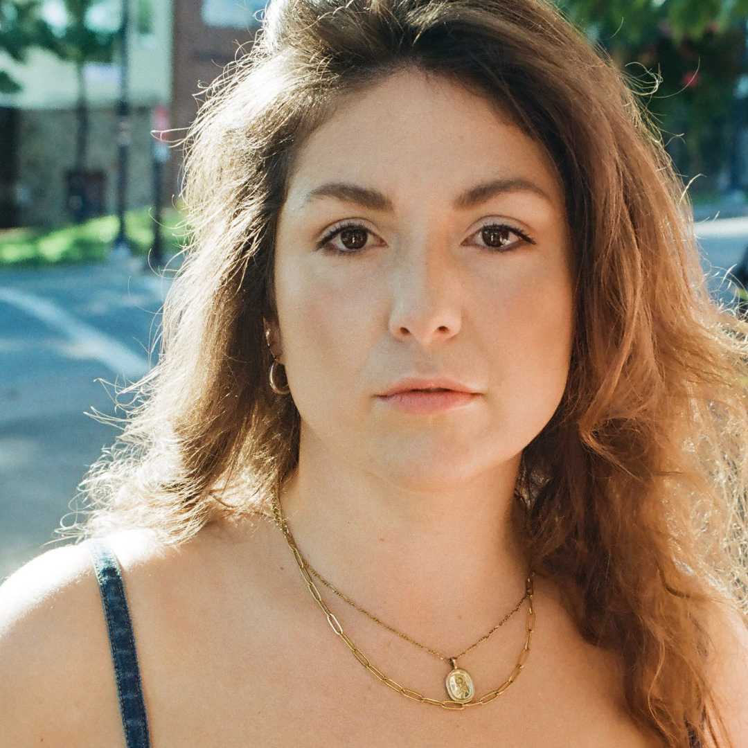 A person with long curly brown hair posing for a headshot. They are wearing a tank top and a gold necklace. 