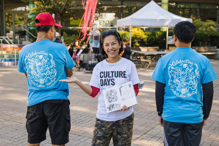 Three people pose for the camera. A woman in the middle faces the camera while wearing a white Culture Days t-shirt while the two others stand on either side of her facing away.