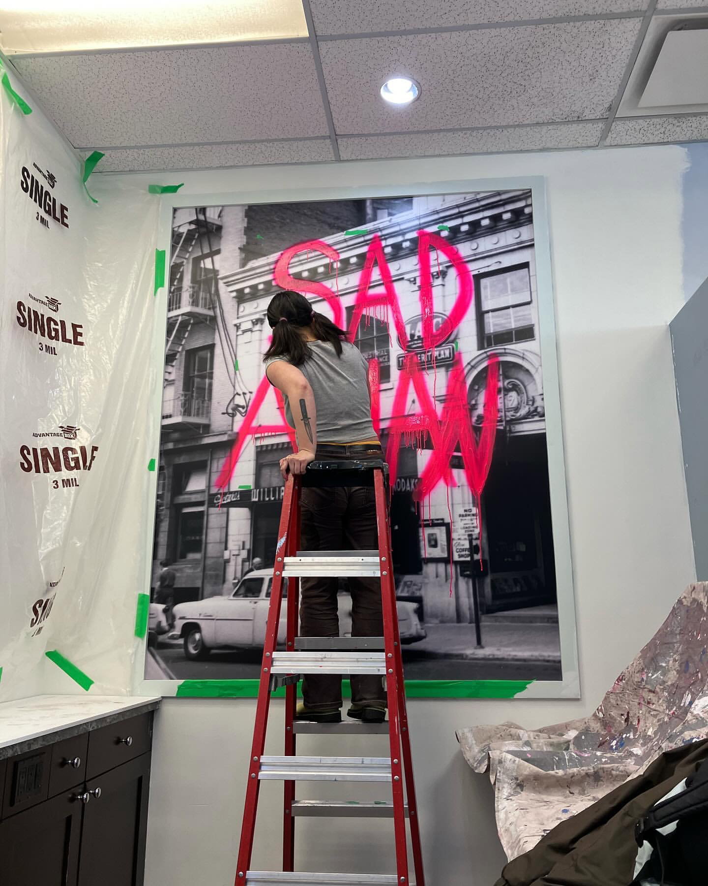 An artist on a ladder, installing a large scale photographic artwork with pink spray painted text that says "Sad Asian."