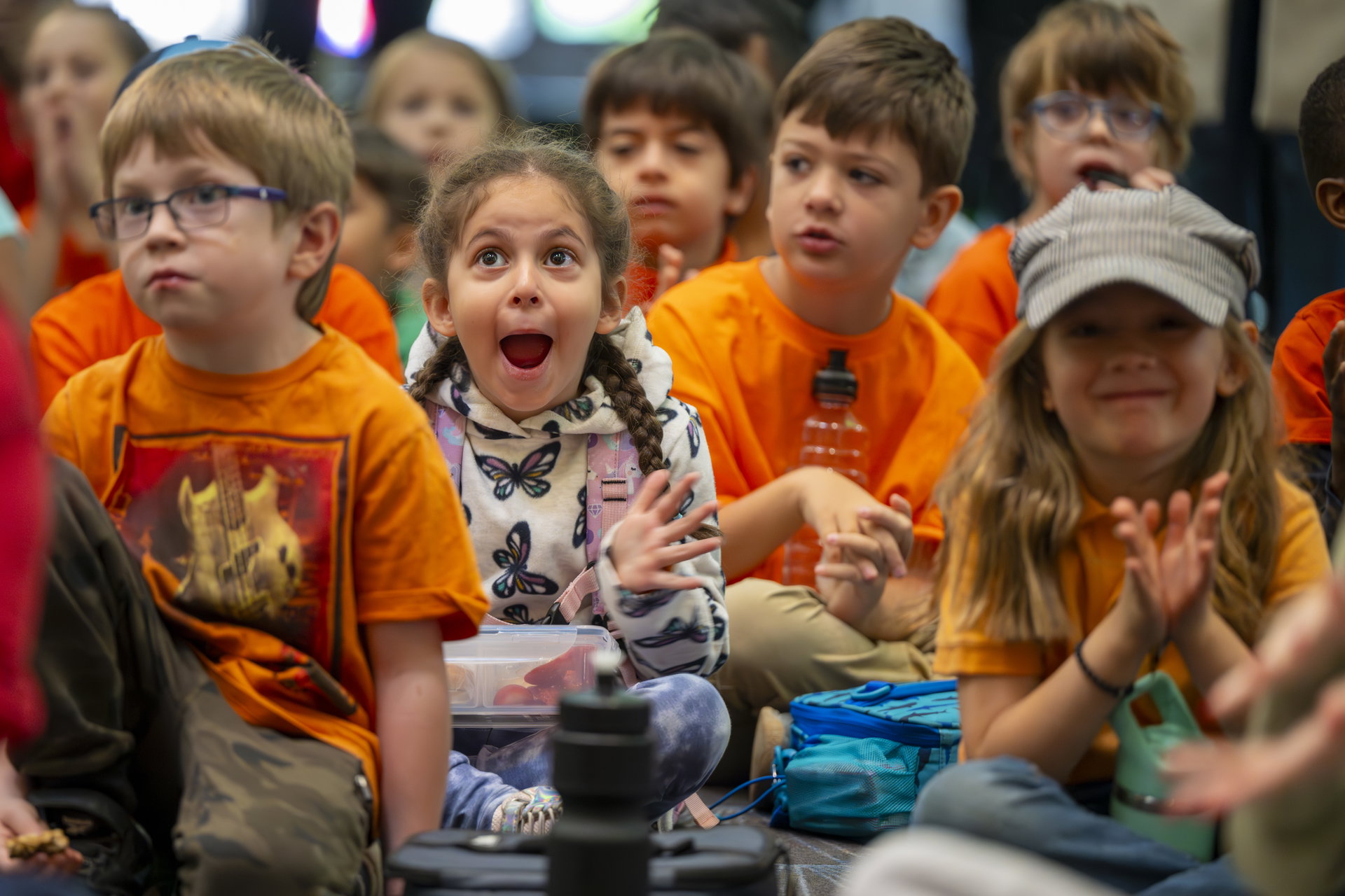 A group of children sit together, some clapping and others watching attentively. One child in the center is excited, with an open-mouthed expression of awe. Many of the children are wearing bright orange shirts.