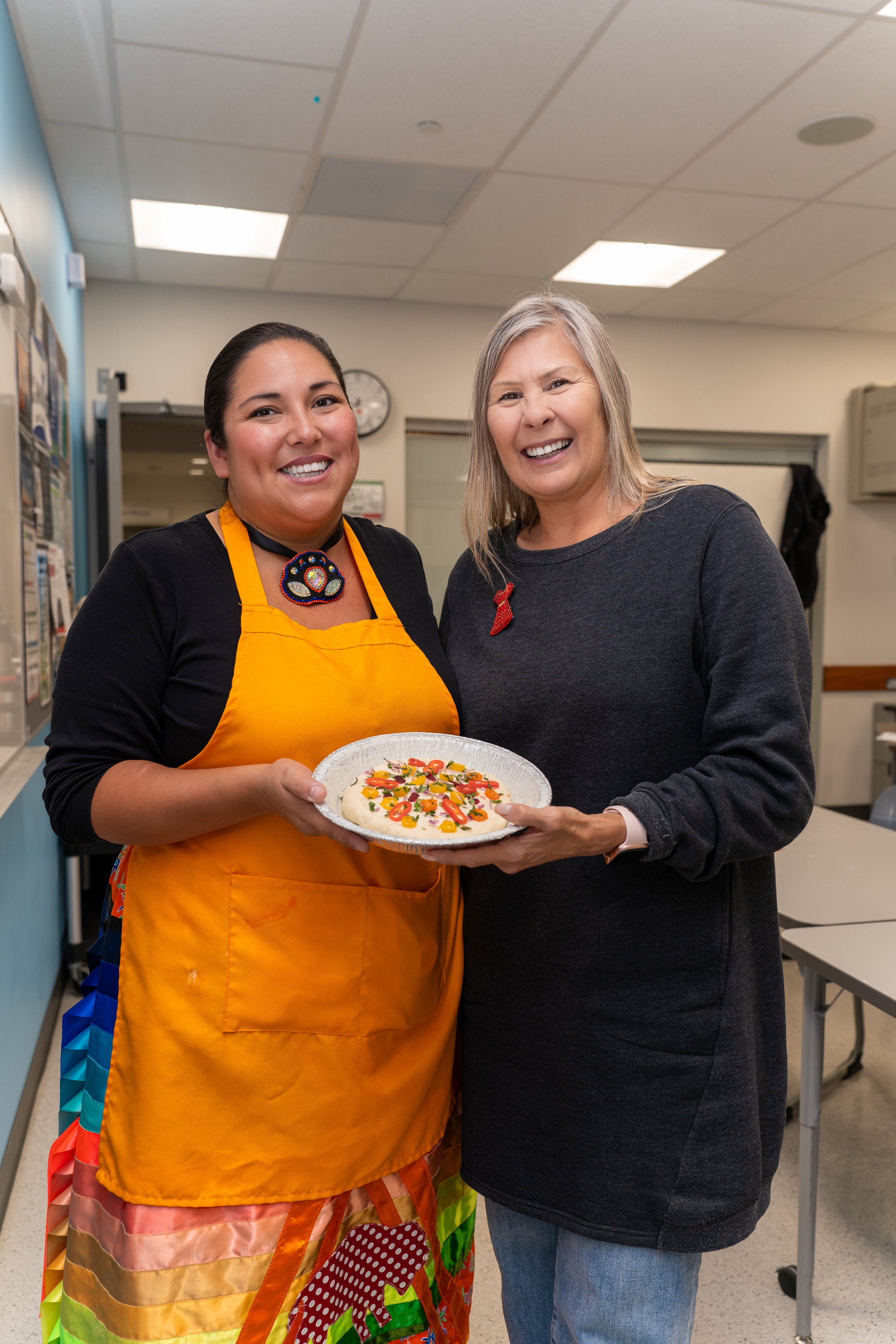 Two smiling individuals stand together in a kitchen, holding a plate of freshly prepared bannock topped with colorful vegetables. One wears an orange apron and traditional ribbon skirt, while the other wears a gray sweater.