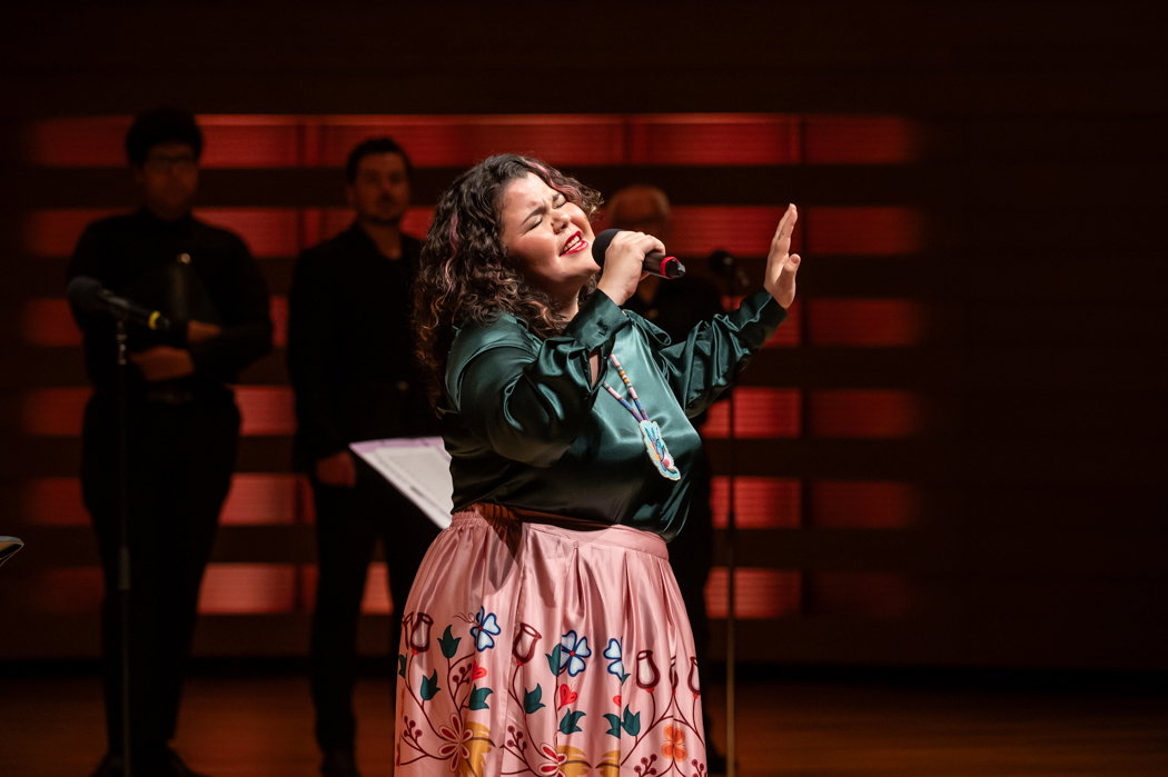 A performer passionately sings into a microphone on stage, wearing a colorful ribbon skirt and a beaded necklace. The background features other performers standing in shadow. The lighting creates a warm, intimate atmosphere.