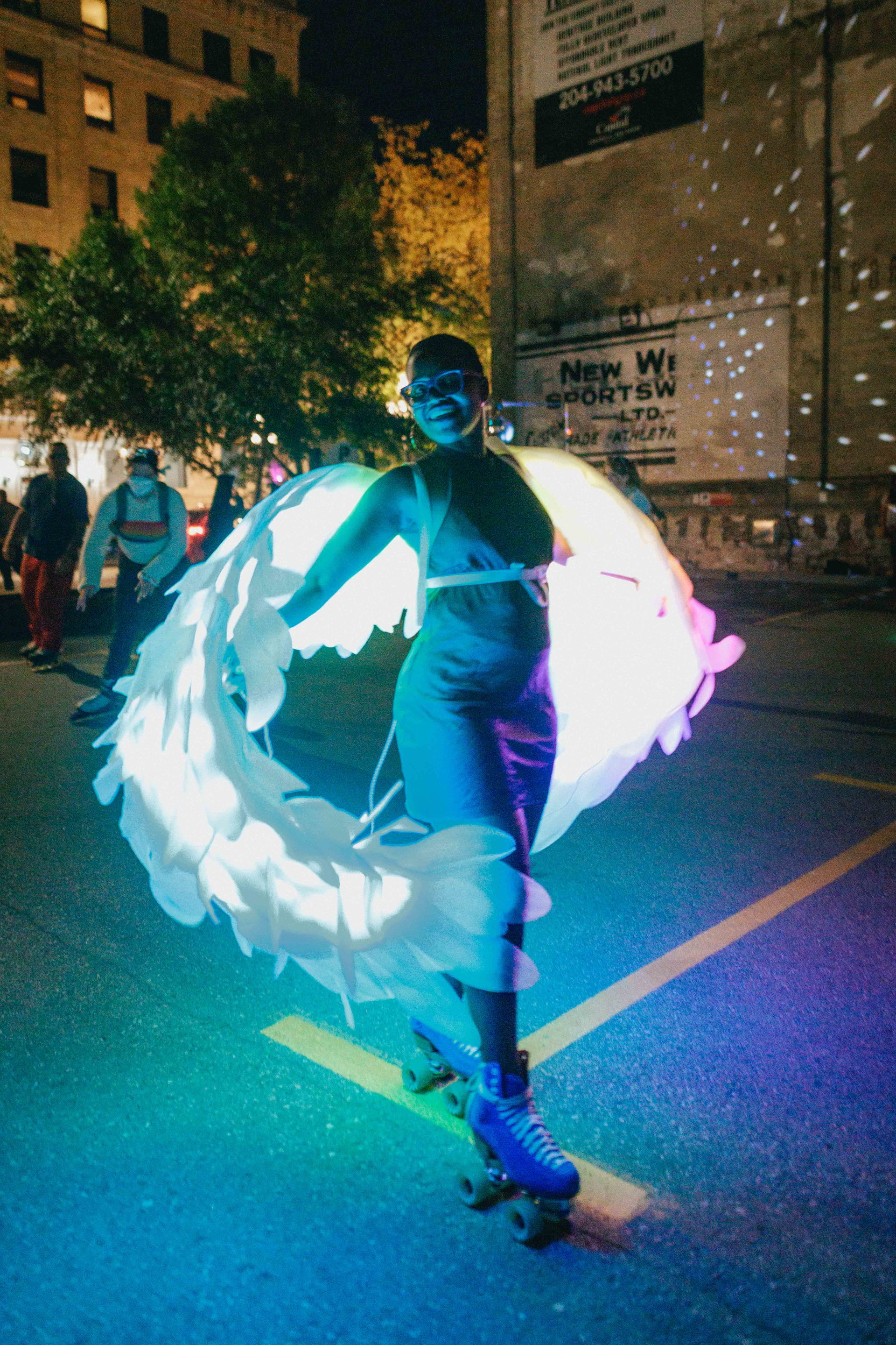 A person on roller skates is illuminated by glowing, wing-like fabric, creating a colorful light display at night. The street scene is vibrant, with people in the background and trees lit by the surrounding glow, giving a festive atmosphere.