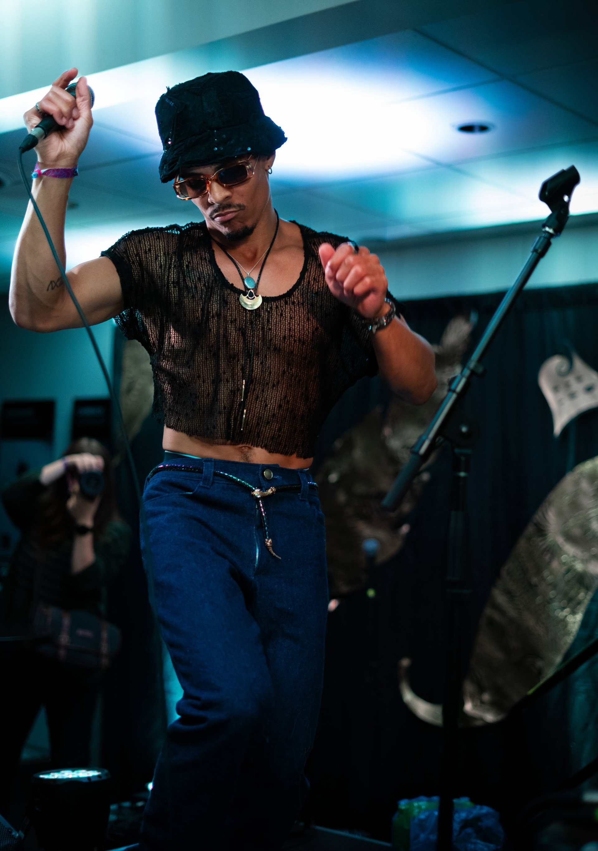 A performer dances on stage, holding a microphone in one hand and wearing sunglasses, a black mesh top, and a bucket hat. The scene is vibrant, with dynamic lighting and a lively atmosphere. A photographer in the background captures the moment.