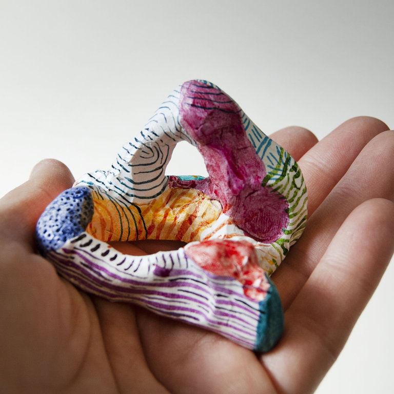 A hand holding a uniquely patterned sculpture. The sculpture has various colors and designs, including red, green, and yellow lines and shapes. The patterns appear to be hand-painted with intricate details on a white background of the sculpture. The image is focused on the sculpture with the hand serving as a scale reference, showcasing the size of the sculpture relative to a human hand.