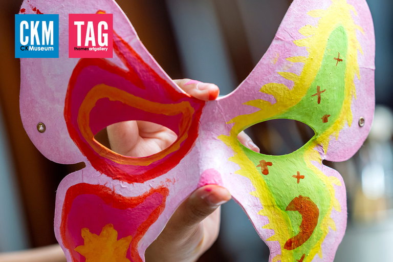 A hand holding a pink mask that is shaped like a butterfly. One side is painted in shades of red and orange, the other side is greens and yellows. Logos for the Chatham-Kent Museum and Thames Art Gallery are found in the top left corner of the image.