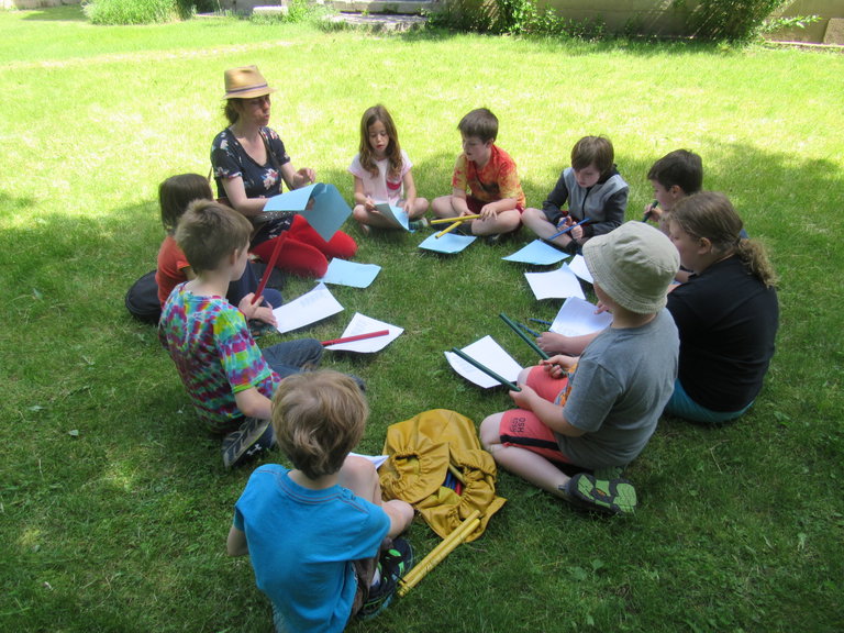 Children are sitting in a circle outside in WAHC's backyard. Several kids have drumsticks and are learning different rhythms from an instructor in the circle.