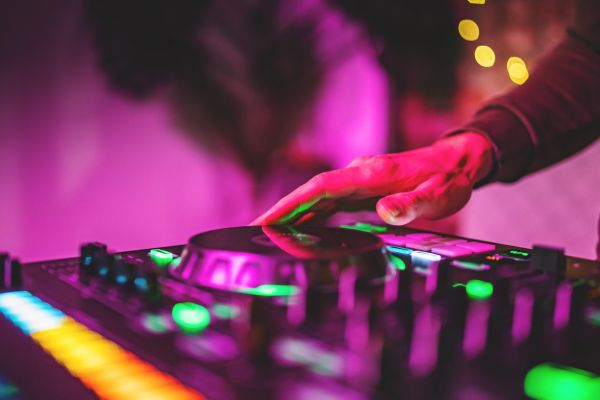 Close-up of a DJ’s hand on a glowing jog wheel of a colourful DJ controller, with multicoloured buttons and knobs lit up against a blurred pink and purple club background.