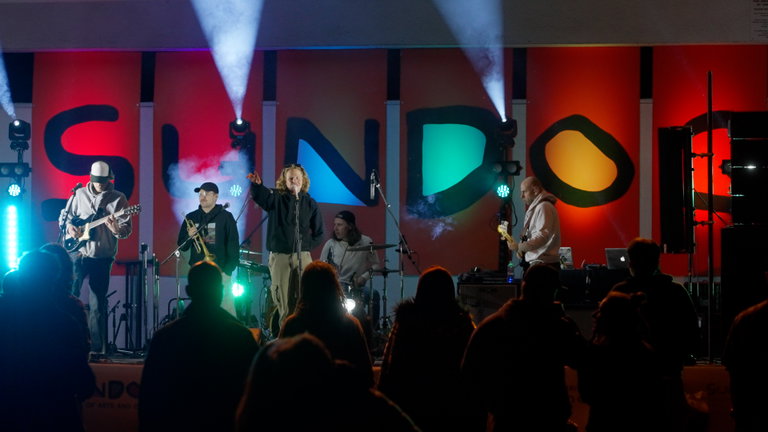 A band plays at the Sundog Festival with large letters spelling SUNDOG behind them. A crowd is watching them and dancing.