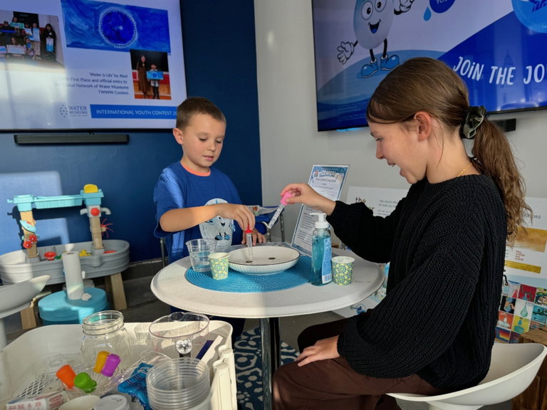 Two people use droppers during a hands-on water science activity at a table inside an educational exhibit space, with water-themed displays and learning materials visible in the background.