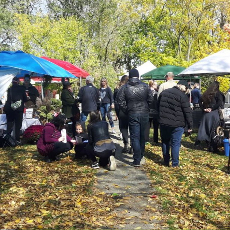 An autumn outdoor market with people walking and browsing various vendor stalls under tents in blue, red, and green. The ground is scattered with fallen leaves, and trees in the background display green and yellow foliage. A few individuals are crouched or bending down, possibly engaging with items on the ground.