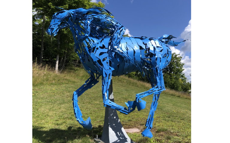 A bright blue sculpture of a horse, made from cut metal pieces, stands in a dynamic running pose on a grassy hill. A cree woman is riding on the horse. Trees with autumn leaves and a cloudy sky are in the background.