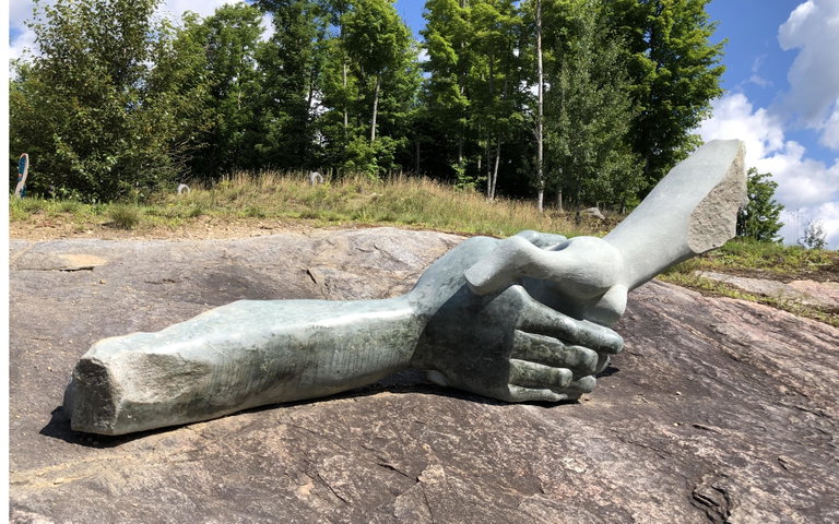 A large sculpture of two hands, carved from light green opal stone, clasped in a handshake. It rests on a flat, dark rock surface with a forest of bare trees and evergreens in the background.