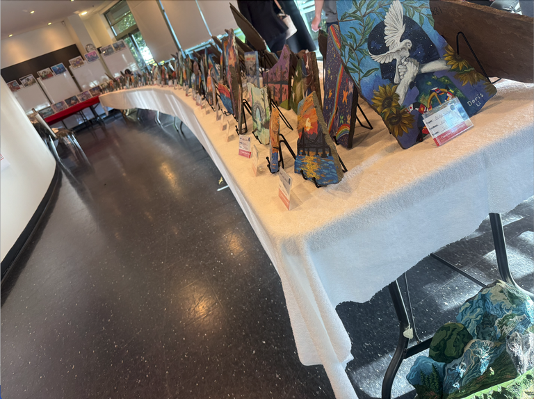 A wide view of a rock painting exhibition display, featuring rows of hand-painted stones arranged on a long table in a gallery space.