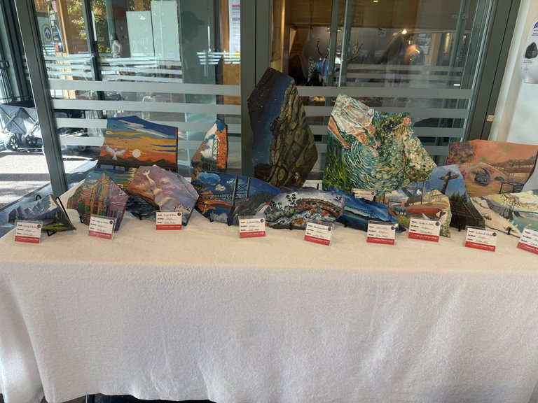 Rock painting exhibition display featuring hand-painted stones on a table, each showing landscapes, cultural scenes, and artistic interpretations of Canada