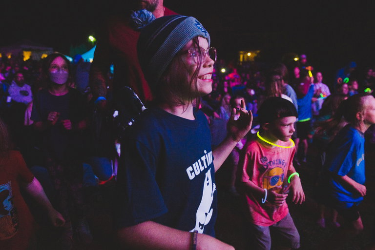 A smiling child wearing a winter toque and Cultivate T-shirt dances in front of the stage during an evening concert, surrounded by families enjoying live music under colourful lights.
