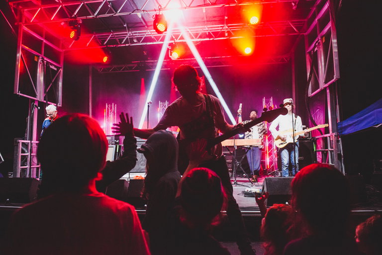 A musician reaches down to high-five children gathered at the front of the stage during a nighttime performance as bright red and white stage lights illuminate the band.