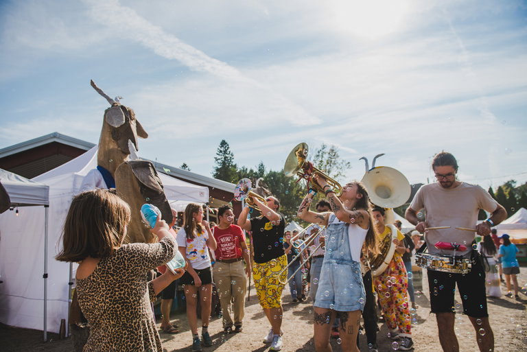 A lively brass band performs while walking through the festival grounds as children, families, and stilt walkers follow along, with bubbles floating through the sunny afternoon.