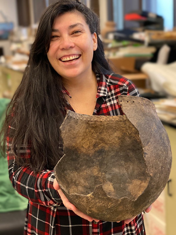 a smiling woman with long, dark hair holds a large, rounded ceramic vessel with dimples along its rim and a large break in its side.