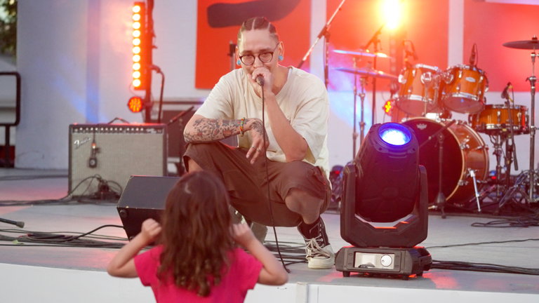 Indigenous rap artist Ray the Nihilist performs at the Sundog Festival of Arts and Culture. He is crouched down on the stage and a young girl dances on the grass below him.