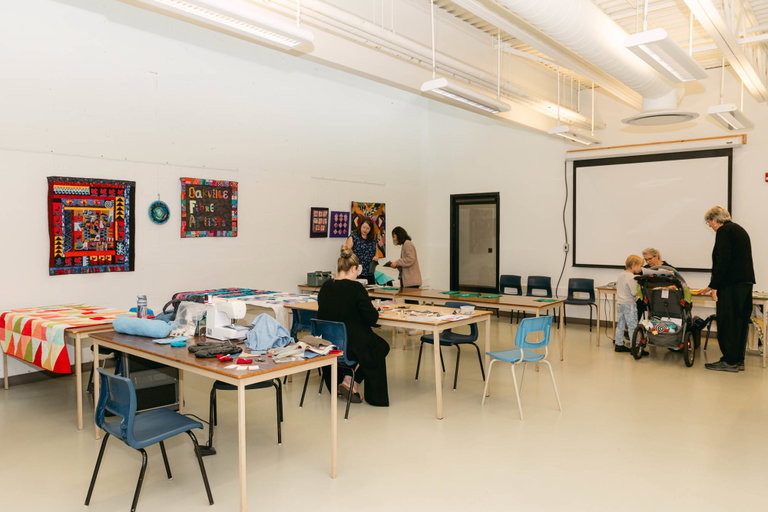 The fibre arts studio set-up with tables and chairs. Participants are looking at displays of sewing materials and quilts on the walls.