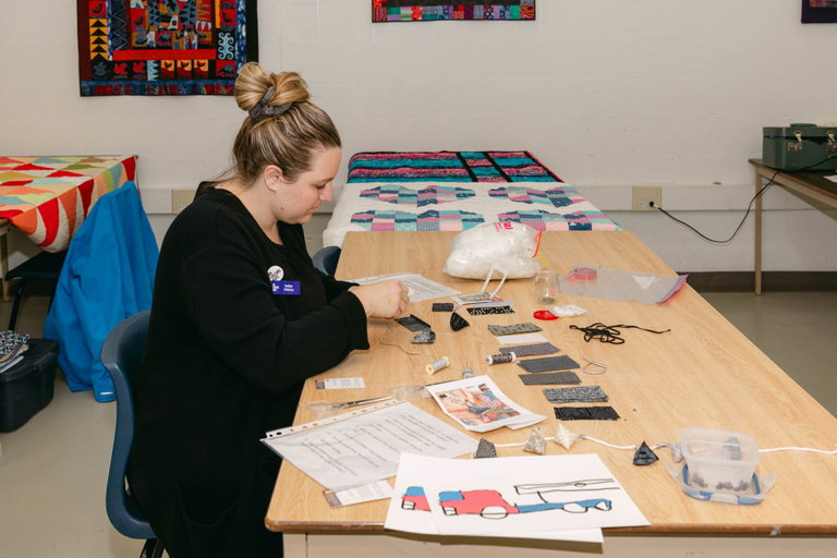 A woman wearing all black, sitting at a wooden table and holding a sewing needle with thread.