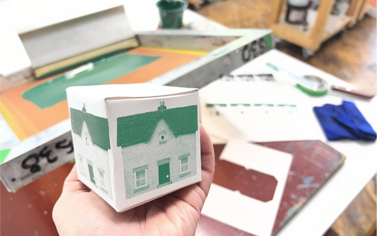 A hand is holding a folded paper cube with the image of a green house printed on two visible sides. The screenprinting setup of how this object was printed could be seen in the background.