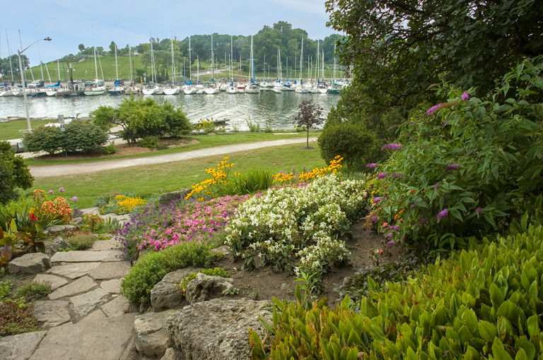 View of rock garden with perennial flowers in bloom. A creek and harbour with sailboats is seen in the distance