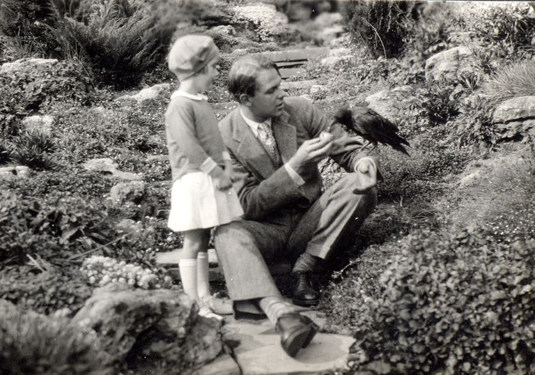 Black and white historic photo of a man sitting on stone steps in a rock garden. A young girl is standing on the step next to him. A crow is perched on the man's arm eating a fruit in the man's hand..