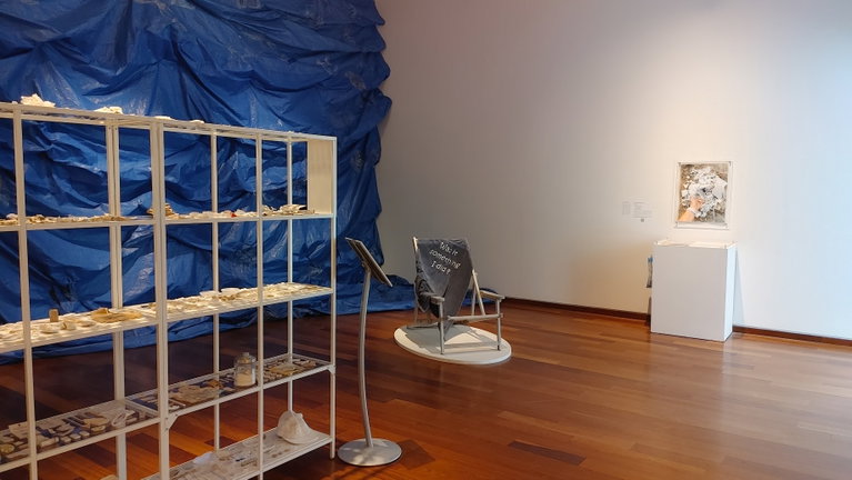 A large installation of white shelves topped with white trash, a large scale blue tarp hung on the wall mimicking the movement of waves, an embroidered blue towel, and a photograph of white trash being piled on the sand. One hand reaches into the picture frame to add another piece of garbage to the pile of refuse which includes paper, plastics, textiles, Styrofoam, cutlery...