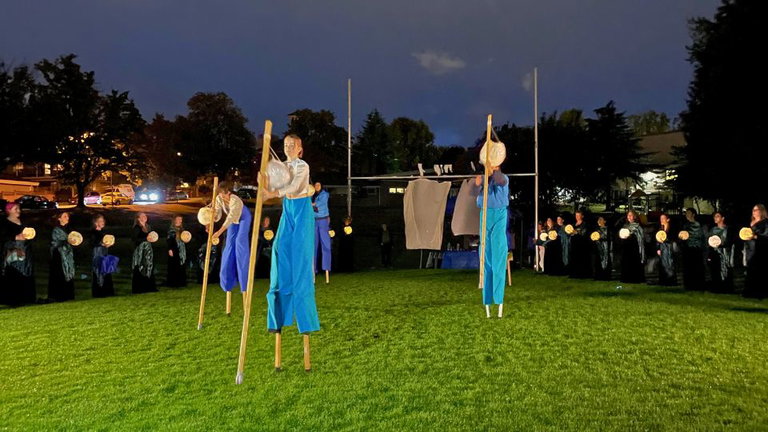 Community performers on stilts with lanterns in a football field park. in the back are women in black with lanterns.