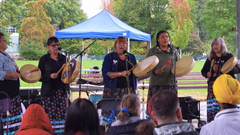 Several women drumming and singing for a crowd in a park.