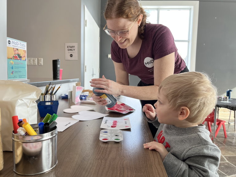 A child stands at a table with an adult pouring paint