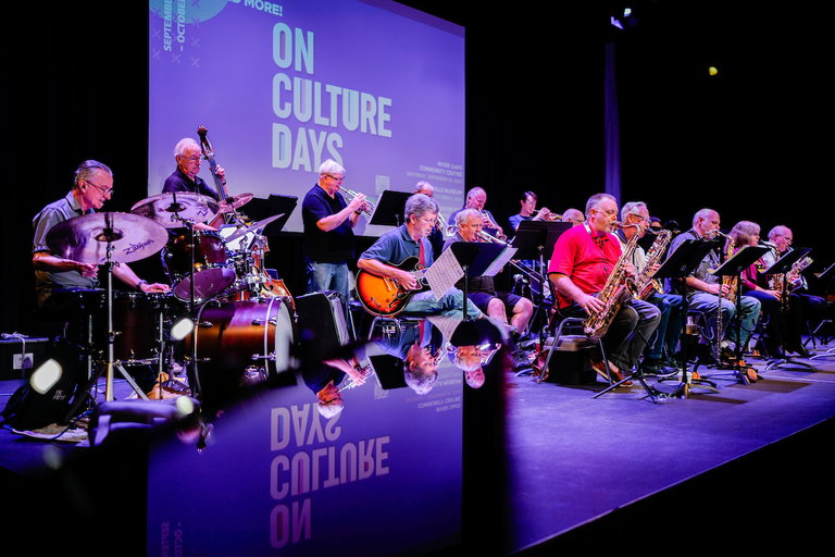 Three rows of musicians from Kerr Street Big Band performing on the Black Box Theatre stage. There's a drummer in the left corner, a guitarist in the centre stage, and saxophones on the right side of the stage.