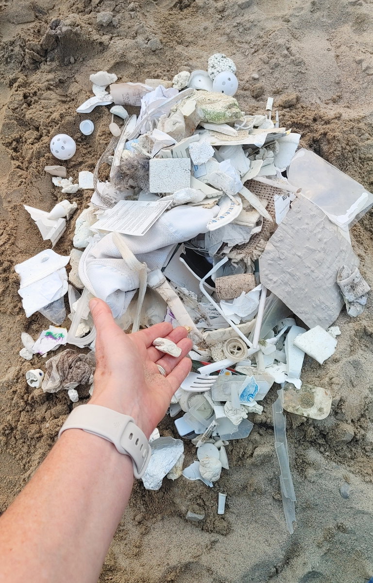An assortment of white trash being piled on the sand. One hand reaches into the picture frame to add another piece of garbage to the pile of refuse which includes paper, plastics, textiles, Styrofoam, cutlery...