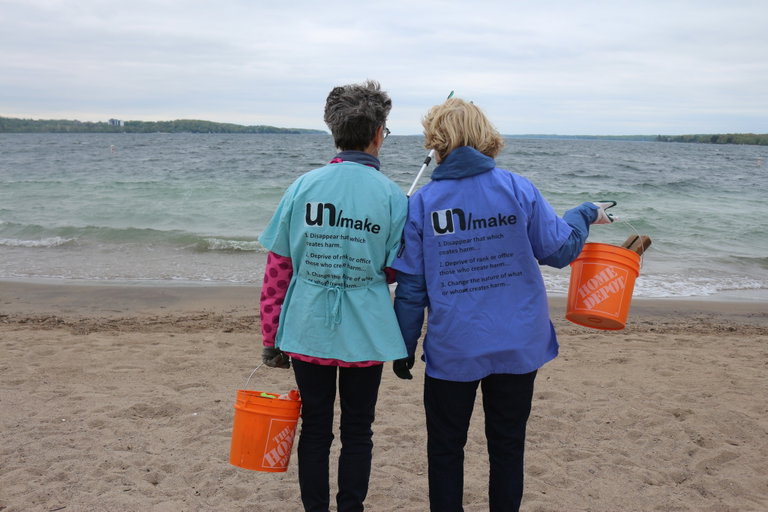 two women standing on a beach wearing UN/making Network beachclean shirts holding buckets full of trash and looking out towards Lake Simcoeing out at Lake Simcoe fro